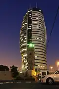 البرج كما يُشاهد ليلاً، يظهر بالصورة أيضاً مئذنة مسجد الجرينة.