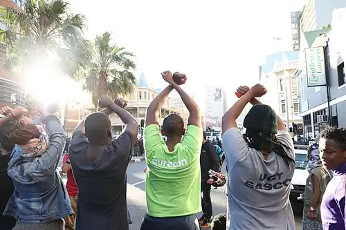 مجموعة من الطلاب يرفعون أيديهم في الهواء للإشارة إلى أنهم جاؤوا بسلام. احتجاج طلابي (2015-16) في جنوب إفريقيا ضد زيادة الرسوم الجامعية.