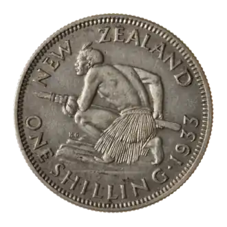 A coin featuring a crouched Māori atop a rocky escarpment. He holds a taiaha war-staff with both hands pointing it and facing forward off the escarpment. He is muscular and shirtless, but wears a piupiu skirt.