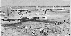 Four 4-engined World War II-era aircraft sitting on the ground at an airstrip. Groups of people are working near each aircraft.