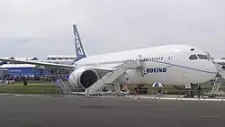 منظر أمامي/جانبي ل787 بيضاء في مكان مكشوف. وضعت السلالم أما المحرك الأيمن للولوج إلى كابينة الطائرة.