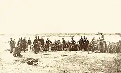 Photograph of a line of uniformed soldiers ranged along a river bank in the foreground with boats on the river in the background