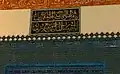 (اضغط للتكبير) اللوحة المكتوبة فوق "المقصورة السلطانية" والتي تذكر اسم مصمم الزخارف بالجامع وسنة انتهاء أعمال الزخرفة: "قد تم نقش هذه العمارة الشريفة بيد أفقر الناس علي بن إلياس علي""في أواخر رمضـــان المبارك سنــة سبع وعشــــرون وثمانمائـــة"