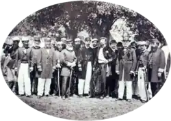 A photograph showing a large group men, most of whom wear uniforms, standing beneath shade trees