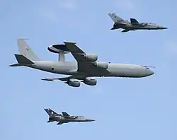 Four-engined jet aircraft with disc-shaped radar on fuselage in-flight flanked by two jet fighters.