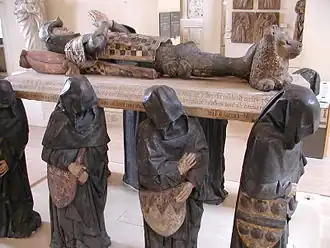 A large sculpture of six life-sized black-cloaked men, their faces obscured by their hoods, carrying a slab upon which lies the supine effigy of a knight, with hands folded together in prayer. His head rests on a pillow, and his feet on a small reclining lion.