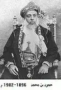 A black-and-white photograph of a man with a white beard wearing a turban, a dark jacket, a white shirt, and a belt and sitting on a chair