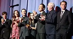 A man in a black suit, a woman in a pink dress, a man in a plaid suit, a woman in a black dress, a Japanese man in a black suit, and an old man in a blue suit clap their hands, while a man in a black suit stands. A microphone stand is in the foreground, and blue curtains are in the background.