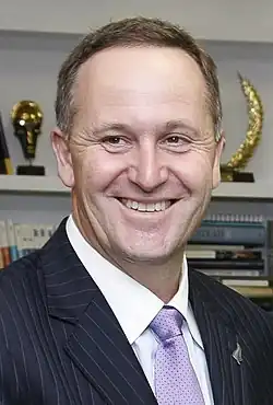 A man with brown eyes and short, brown hair wearing a white shirt, purple tie, and a black jacket with white pin-stripes.