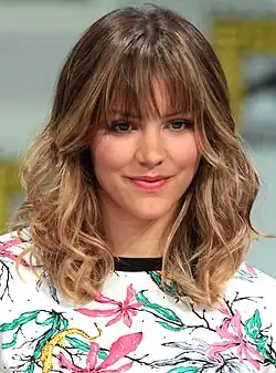 Katharine McPhee is pictured outdoors with her right hand up slightly waving with a small smile not showing teeth; Her long dark hair with lightened tips is up in a messy bun. She is wearing a little black dress with a plunging neck line.