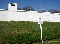A tall white-painted wall with an overlapping round top and a square turret on the left-hand side. Behind it is a grey watchtower with an octagonal top and a searchlight on the roof. Further back are a group of white-painted half-timbered buildings and several trees.