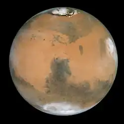 An orange disk with a darker region at center and darker bands in the upper and lower halves. A white patch at the top is an ice cap, and fuzzy white regions at the bottom and the right side of the disk are cloud formations.