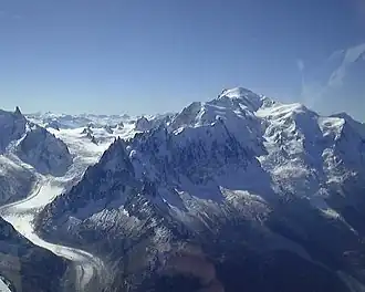 Photograph of a towering snow-covered mountain at right, a glacier at left, and a clear blue sky.