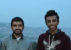 Two young men, one in a collared shirt and one in a maroon hoodie, facing the camera