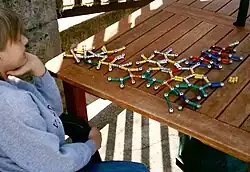 Seated boy facing 3/4 away from camera, looking at a ball-and-stick model of a molecular structure. The model is made of colored magnets and steel balls.