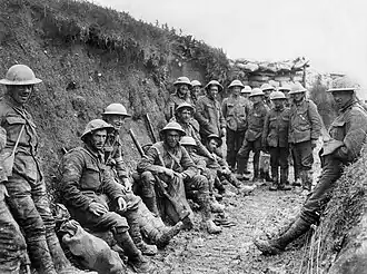 Black and white photo of two dozen men in military uniforms and metal helmets sitting or standing in a muddy trench.