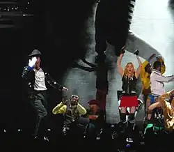 A group of males and a female performing on stage in front of a crowd of people. A male is shown blowing a kiss to the audience while wearing a white glove in his right hand and a white shirt with black pants and a jacket. An African American male wearing a white and dark grey shirt is shown in a crouched-down position. The female is waving her hands in the air while wearing black fingerless gloves with a black shirt and pink shorts. There are also two other African American males in the background making hand gestures. Behind the people on the stage, there is a screen that shows a black and white photo.