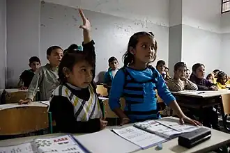 Children at desks in a classroom. &nbsp;One child raises her hand.