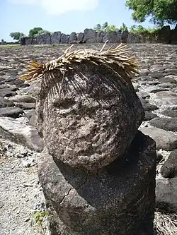 Weathered grey stone bust with a palm frond crown. Grey rocks with white and in between them in the background.