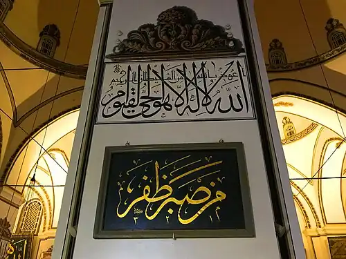أعوذ بالله من الشيطان الرجيم ﴿اللَّهُ لَا إِلَٰهَ إِلَّا هُوَ الْحَيُّ الْقَيُّومُ﴾ - وتحتها لوحة مُعلقة كُتب عليها: «من صَبَرَ ظَفَرَ»