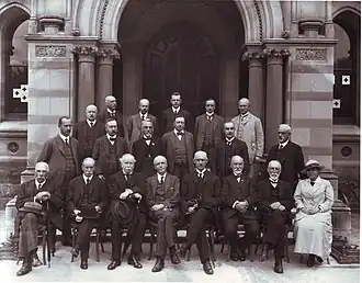 A group of people pose for a formal group portrait beneath a stone arch. There is only one woman in the group. While the men have removed their hats, she still wears hers.المجلس الاستشاري لجامعة أديليد في عام 1919. وتبدو هيلين مايو الأولى من اليمين في الصف الأول.