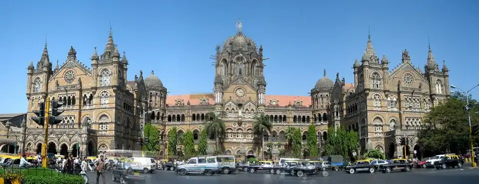 A brown building with clock towers, domes and pyramidal tops. Also a busiest railway station in India. A wide street in front of it